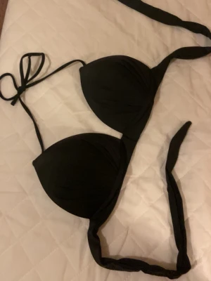 Svart bikiniöverdel med knyt - Stilren svart bikiniöverdel med vadderade kupor och knyt i nacken och ryggen. Klassisk trekantsmodell som ger en snygg siluett och passar perfekt till sommaren. Materialet känns mjukt och följsamt mot huden.