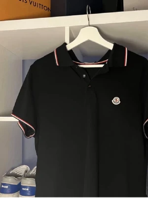 Svart Moncler pikétröja - Snygg svart pikétröja från Moncler med klassisk krage och knappar framtill. Tröjan har vita och röda detaljer på krage och ärmslut samt Moncler-logga på bröstet. Perfekt för en stilren och sportig look.