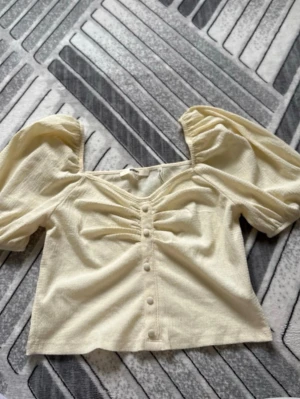 Ljusgul topp med puffärm och knappar - Trendig ljusgul topp med puffärmar och dekorativa knappar framtill. Toppen har en lätt croppad passform och rynkade detaljer vid bysten. Perfekt för dig som vill ha en söt och stilren look.