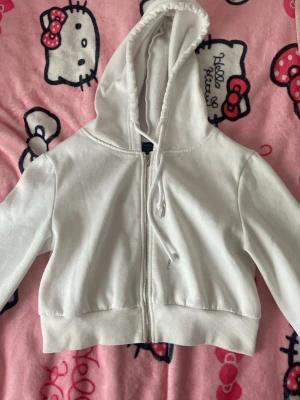 Vit croppad hoodie med dragkedja - Säljer en vit croppad hoodie med dragkedja framtill och justerbar huva med snören. Hoodien har breda muddar vid ärmslut och nederkant. Perfekt till chill outfits och enkel att matcha med jeans eller mjukisbyxor. Hoodien har en väldigt liten brun fläck i mitten som finns i bild, syns knappt, men annars så är den i bra skick!