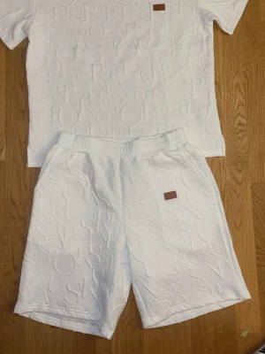 Vitt set med reliefmönster - Säljer ett vitt set med t-shirt och shorts i matchande material. Båda plaggen har ett upphöjt reliefmönster med bokstäver och en liten brun patch framtill. Setet har en avslappnad passform och resår i midjan på shortsen. Perfekt för en clean och modern look.