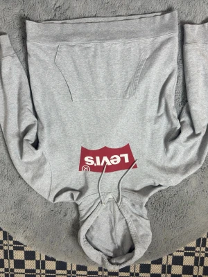 Grå Levi's hoodie med logga - Snygg grå hoodie från Levi's med stor röd logga på bröstet. Klassisk känguruficka framtill och justerbar huva med dragsko. Tillverkad i mjukt bomullsmaterial som är perfekt för en avslappnad stil.