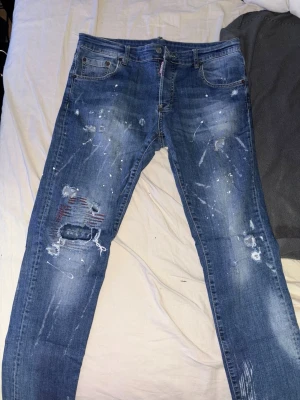 Dsquared2 Jeans Storlek 50 - Dsquared2 Jeans, storlek 50,  blå jeans med distressed detaljer, hål och färgstänk för en riktigt edgy look. Jeansen har raka ben, fem fickor och coola slitningar på både fram- och baksida. Perfekta för dig som gillar streetwear och vill sticka ut.