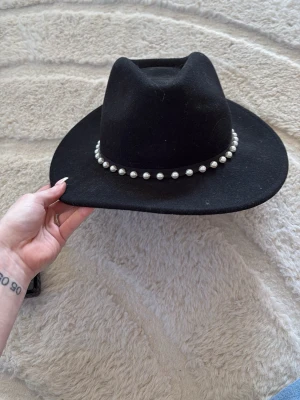 Svart cowboyhatt med pärldetaljer - Snygg svart cowboyhatt med bred brätte och dekorativt band med vita pärlor runt kronan. Hatten har en klassisk form och är tillverkad i filtmaterial som ger en cool och trendig look.