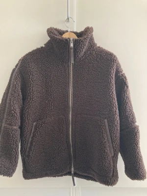 Mörkbrun teddyjacka med dragkedja - Mysig mörkbrun teddyjacka med hög krage och hel dragkedja framtill. Jackan har två stora fickor och är gjord i fluffigt, mjukt material som ger en riktigt skön vibe. Perfekt för kyliga dagar när du vill vara både varm och stylish.