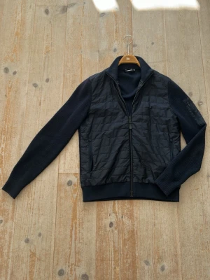 Marinblå hybridtröja från J.Lindberg - Snygg marinblå hybridtröja från J.Lindberg med quiltad front och stickade ärmar. Tröjan har dragkedja framtill, hög krage och ribbade muddar. Perfekt för lager-på-lager och ger en sportig men stilren look.