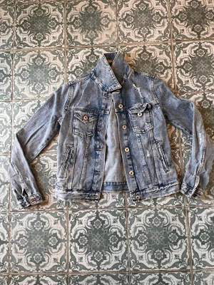 Ljusblå vintage jeansjacka - Säljer en ljusblå jeansjacka med slitna detaljer och klassisk knappstängning. Jackan har två bröstfickor med lock och knappar samt två sidofickor. Snygg retrostil med hög krage och justerbara knappar i midjan. Perfekt för dig som gillar en avslappnad och trendig look.