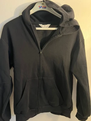 Svart hoodie med dragkedja från H&M - Klassisk svart hoodie från H&M med dragkedja framtill och två stora fickor. Tillverkad i mjuk bomullsblandning och har en rymlig huva. Perfekt för en avslappnad och stilren look.