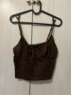 Brun linnetopp från Bik Bok - Snygg brun linnetopp från Bik Bok med tunna axelband och knyt framtill. Toppen har en croppad passform och är tillverkad i ett mjukt, glansigt material som känns skönt mot huden. Perfekt att styla med jeans eller kjol för en trendig look.