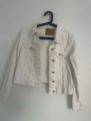 Vit jeansjacka  - Snygg vit jeansjacka med klassisk krage, långa ärmar och silverfärgade knappar framtill. Två bröstfickor med lock och knappdetaljer ger en tidlös look. 