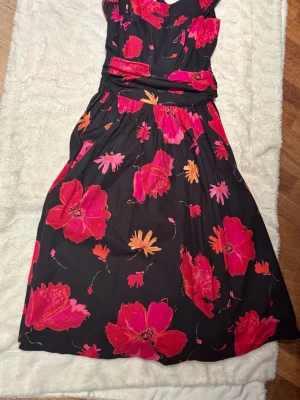 Svart klänning med stora blommor. Laura Ashley - Svart klänning med stora, färgstarka blommor i rosa, rött och orange. Klänningen har en markerad midja och v-ringad urringning. Kjolen är vid och faller snyggt. Perfekt för dig som vill sticka ut med ett färgglatt mönster.