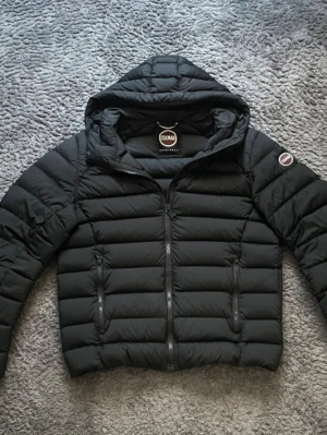 Svart dunjacka med huva från Colmar - Snygg svart dunjacka från Colmar med huva och två dragkedjeförsedda fickor framtill. Jackan har quiltad design och en Colmar-logga på ärmen. Perfekt för kalla vinterdagar och riktigt skön att bära.
