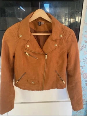 Brun skinnjacka med gulddetaljer - Snygg brun skinnjacka från H&M i bikerstil med guldfärgade dragkedjor och knappar. Jackan har tre fickor framtill och en cool asymmetrisk dragkedja. Perfekt för dig som vill ha en trendig och stilren look.