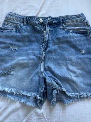 Blå denimshorts med fransig kant - Säljer ett par blå denimshorts med hög midja och fransig kant. Shortsen har slitna detaljer framtill, klassiska fickor och dragkedja med knapp. Perfekta för sommaren och ger en avslappnad vibe.