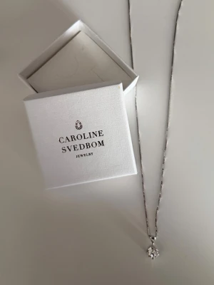 Silverhalsband med klar Swarovski sten Caroline Svedbom - Classic Petite Necklace är ett klassiskt halsband med en gnistrande swarovskikristall. Den vackra venetzialänken är av sterling silver 925 med 18K guld- eller rhodiumplätering. Kedjan är ca 50 cm lång och knäpps med fjäderlås. Mått 8 X 10 mm. Designad i Sverige, handgjord i Grekland. Levereras i Caroline Svedbom ask.  Nyskick!