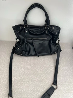 Svart handväska med nitar och dragkedja - Snygg svart handväska i konstläder med silverfärgade nitar och detaljer. Väskan har både handtag och en lång axelrem, samt en praktisk dragkedjeficka framtill. Perfekt för dig som gillar en edgy och stilren look.