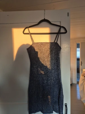 Glittrig mörkblå kort klänning - Supersnygg mörkblå kort klänning med tunna axelband och glittrigt tyg. Klänningen har en figurnära passform och är tillverkad i ett stretchigt syntetmaterial som sitter bekvämt på kroppen.