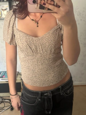 Beige prickig blus med puffärm - Supersöt beige blus med svarta prickar och puffärmar. Toppen har smockad midja, v-ringad urringning och korta ärmar. Perfekt för dig som gillar en feminin och trendig stil.