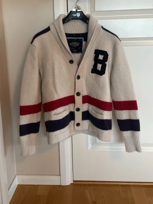 Vit kofta från Bondelid med ränder - Säljer en vit kofta från Bondelid med stora knappar, breda röda och marinblå ränder samt ett stort B på bröstet. Storleken står M men sitter mer som S eller Xs