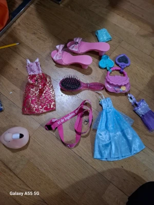 Docktillbehör: kläder, skor och accessoarer - Ett set med docktillbehör som inkluderar två klänningar (en röd med guldmönster och en blå glittrig), ett par rosa klackskor, en rosa borste, en rosa handväska, ett blått plånboksliknande tillbehör och ett rosa nyckelband. Perfekt för att styla dockor.