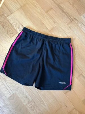 Svarta träningsshorts med rosa detaljer - Svarta träningsshorts från IN-MOTION med rosa ränder längs sidorna. De har elastisk midja och är tillverkade i ett lätt och syntetiskt material som passar perfekt för träning eller sportiga aktiviteter.