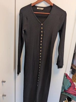 Svart ribbad långklänning Envii - Svart långklänning från Envii med ribbad struktur och v-ringning. Klänningen har långa ärmar och knappar hela vägen framtill, vilket ger en snygg detalj. Perfekt för dig som gillar en stilren och enkel look.