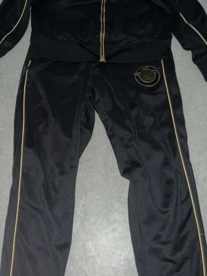 Svart EA7 loungewear-set med guld. PRIS KAN DISKUTERAS! - Snyggt svart loungewear-set från EA7 med guldfärgade detaljer och dragkedja. Setet har guldiga ränder längs benen och EA7-logga på byxbenet samt muddar med EA7-tryck. Perfekt för chill dagar hemma eller när du vill vara bekväm men ändå se fräsch ut.