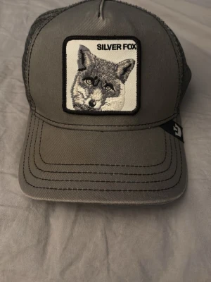 Grå truckerkeps Silver Fox Goorin Bros - Snygg grå truckerkeps från Goorin Bros med broderad patch framtill med en räv och texten 'SILVER FOX'. Kepsen har böjd skärm, mesh baktill för ventilation och justerbar snapback. Perfekt för en avslappnad stil nu till sommaren. Är köpt på hatstore. Tveka inte på att höra av dig vid minsta lilla, pris kan diskuteras!//