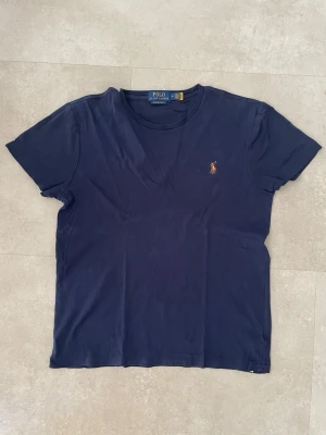 Mörkblå t-shirt från Polo Ralph Lauren - Klassisk mörkblå t-shirt från Polo Ralph Lauren med korta ärmar och den ikoniska broderade loggan på bröstet. Tillverkad i mjuk bomull för en skön känsla och perfekt passform. Enkel och stilren design som funkar till det mesta.