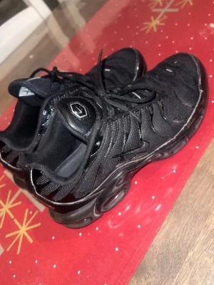 Nike Air Max Plus Tn helsvarta sneakers - Säljer ett par helsvarta Nike Air Max Plus Tn sneakers med klassisk mesh- och syntetöverdel, svarta snören och ikoniska Tn-detaljer på hälen. Skorna har en chunky sula med synlig dämpning och sportig design som sticker ut.