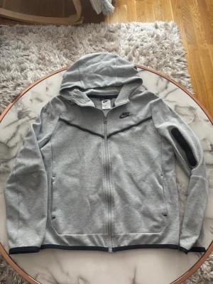 Grå zip hoodie från Nike - Snygg grå hoodie från Nike med dragkedja och svart Nike-logga på bröstet. Hoodien har svarta detaljer längs dragkedjan och på ärmen, samt två fickor framtill. Tillverkad i mjukt material och har en klassisk huva.