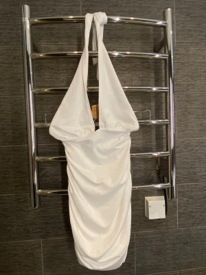 Vit halterneck kortklänning - Snygg vit kort klänning med halterneck och draperad kjol. Klänningen har en djup urringning och är tillverkad i ett glansigt material som ger en lyxig känsla. Perfekt för dig som vill sticka ut med en elegant och trendig look.
