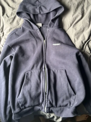 Mörkblå zip hoodie med brodyr - En mörklila hoodie med dragkedja och huva. Den har två stora fickor framtill och en liten vit brodyr på bröstet. Hoodien är i mjukt material och har ribbade muddar vid ärmslut och nederkant. Perfekt för en chill och avslappnad stil.
