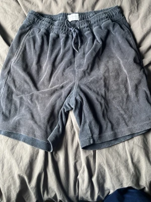 Grå frottéshorts med snörning - Mysiga grå shorts i mjuk frotté med elastisk midja och snörning framtill. Shortsen har en avslappnad passform och en bakficka. Perfekta för sommardagar eller chilla hemma.