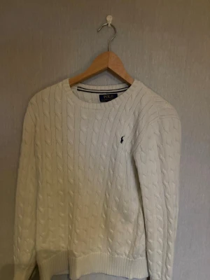 Vit kabelstickad tröja Polo Ralph Lauren - Vit kabelstickad tröja från Polo Ralph Lauren med rund halsringning och broderad logga på bröstet. Tröjan har långa ärmar och ribbstickade muddar vid ärmslut och nederkant. Perfekt för en klassisk och stilren look.