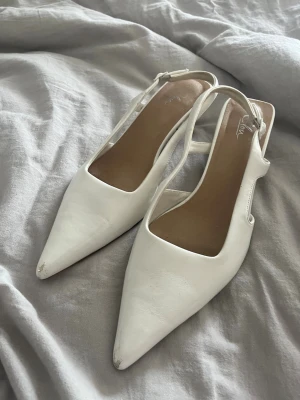 Vita pumps med slingback och klack - Säljer ett par vita pumps från din sko med spetsig tå, slingback-rem och snygga utskärningar på sidorna. Skorna har en medelhög klack och är tillverkade i ett slätt material som ger en stilren look. Perfekta för att lyfta din outfit. Slitage på spetsen från användning, använd 2 gånger