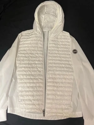Vit pufferjacka från Colmar - Snygg vit pufferjacka från Colmar med quiltad front, huva och dragkedja. Jackan har ett Colmar-märke på ärmen och är tillverkad i lätt, glansigt material. Perfekt för kalla dagar när du vill hålla stilen.