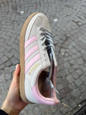 Adidas Spezial sneakers grå/rosa - Snygga Adidas Spezial sneakers i grå mocka med klassiska rosa ränder och detaljer. Skon har en beige gummisula och ljusgrå skosnören. Perfekt för dig som gillar retrostil och vill sticka ut med färg. Ikonisk Adidas-logga i guld på sidan. Finns från storlek 37-40