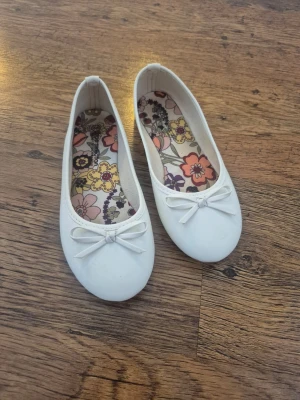 Vita ballerinaskor med rosett - Snygga vita ballerinaskor med rund tå och dekorativ rosett framtill. Skorna har en färgglad blommig innersula. Perfekt till sommaren.💗