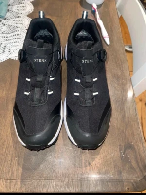 Svarta sneakers från STENK - Svarta sneakers från STENK med vit sula och smart snörningssystem. Skorna är tillverkade i ett vattenavvisande material och har en robust design med detaljer i svart och vitt. Perfekta för dig som vill ha både stil och funktion.