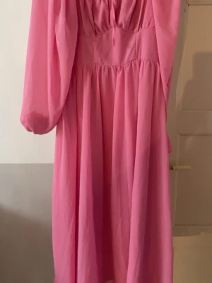 Rosa långklänning med puffärmar - En rosa långklänning med vacker rynkad överdel och långa puffärmar. Klänningen har en luftig passform och faller snyggt ner mot golvet. Perfekt för dig som vill sticka ut med en färgstark och feminin look.