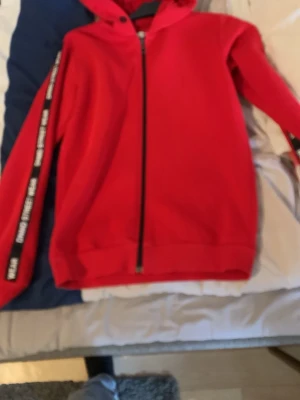 Röd hoodie från DNMD Street Wear - Snygg röd hoodie med dragkedja från DNMD Street Wear. Huva och ribbade muddar. Svarta band med vit text längs ärmarna ger en cool streetstil. Perfekt för dig som vill sticka ut lite extra. Mjuk och skön bomullsblandning.
