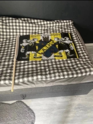 AIK supporterflagga med tryck - AIK supporterflagga i svart, gult och vitt med klubbens emblem och två tecknade figurer. Texten 'Allmänna Idrottsklubben Stockholm' syns tydligt. Flaggan är fäst på en träpinne och passar perfekt för matcher eller supporterträffar.