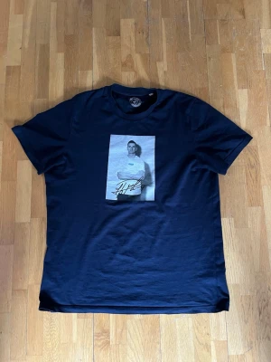 Signerad Haris Radetinac tröja - Svart t-shirt med ett stort tryck framtill föreställande en person i svartvitt och Djurgården-logga på tröjan. Trycket har även en autograf. Klassisk rund hals och korta ärmar. Perfekt för dig som gillar streetwear och sportig stil.