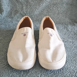 Vita slip-on sneakers från Gant - Stilrena vita slip-on sneakers från Gant med broderad logga framtill. Skorna har en klassisk låg profil och ovandel i canvas, samt bruna gummisulor med mönster och Gant-detaljer undertill. Perfekta för en clean och avslappnad look.