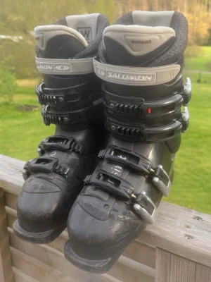 Svarta pjäxor från Salomon - Säljer ett par svarta pjäxor från Salomon med fyra spännen och grå detaljer. De har en robust design och är perfekta för dig som gillar vintersport. Märket syns tydligt på remmen och insidan är vadderad för extra komfort. Bra skick. MÄRKE-SALOMON. STORLEK-41,5. PRIS-350kr.