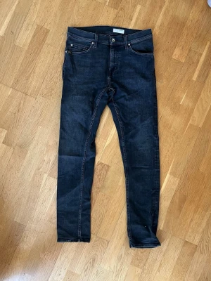 Mörkgrå pistolero jeans från tiger of sweden  - Säljer ett par mörkgrå pistolero jeans från Tiger of Sweden med klassisk femficksdesign och smal passform. Jeansen har orange sömmar och är tillverkade i stretchigt denimtyg för extra komfort. Perfekta för en trendig och avslappnad stil.