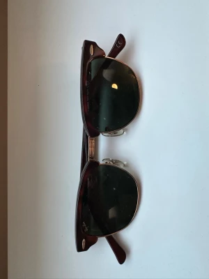Ray-Ban Clubmaster solglasögon brun - Snygga Ray-Ban Clubmaster solglasögon med klassisk brun båge och guldiga detaljer. Glasen är mörkgröna och modellen har en retroinspirerad form med metallbrygga och näskuddar för extra komfort. Perfekt accessoar för en trendig look.