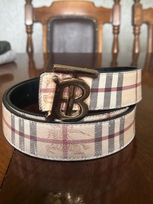 Rutig skärp från Burberry med B-spänne - Snyggt skärp från Burberry med klassiskt rutigt mönster i beige, svart och rött. Skärpet har ett stort metallspänne med Burberrys ikoniska B-logga. Tillverkat i skinn och passar perfekt för att lyfta vilken outfit som helst.