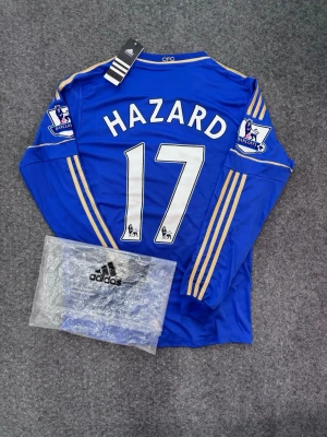 Chelsea Hazard 17 långärmad tröja - Snygg blå Chelsea-tröja från Adidas med Hazard och nummer 17 på ryggen. Långärmad modell med gula detaljer och Premier League-patch på ärmarna. Tillverkad i funktionsmaterial, perfekt för fotboll eller träning. Klassisk Samsung-logga på bröstet.